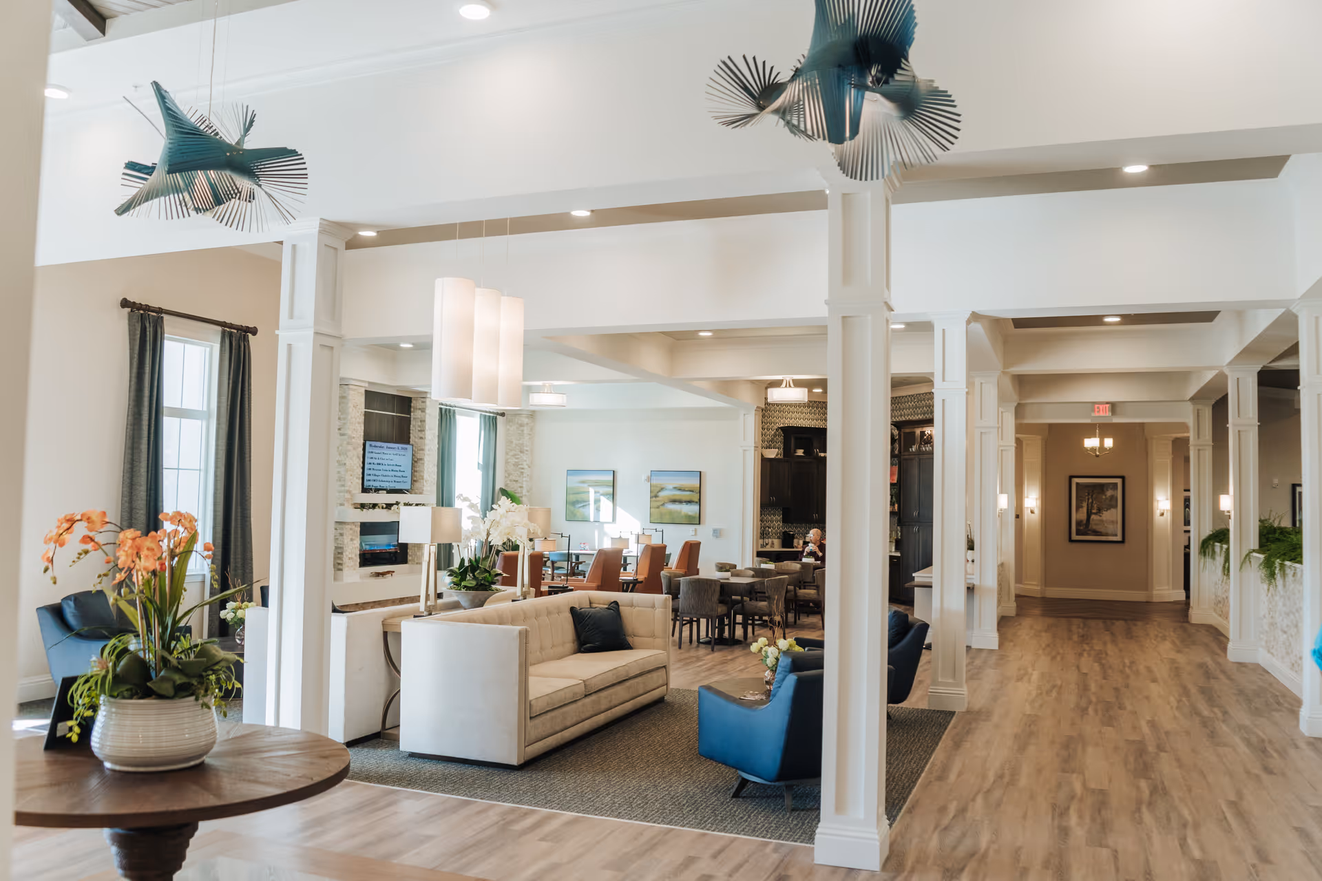 A bright and spacious senior living facility common area with modern decor, featuring a beige sofa, blue armchairs, a round wooden table with a flower arrangement, and a dining area with tables and chairs in the background. The space has white pillars, wood flooring, large windows with curtains, and decorative ceiling lights.