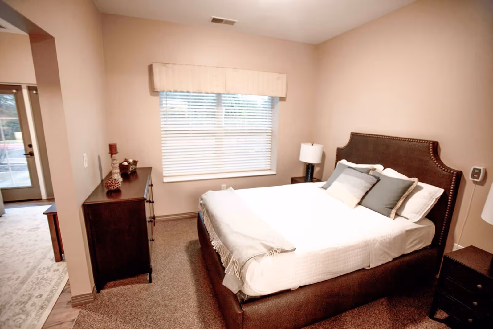 A cozy furnished bedroom with a queen bed, upholstered headboard, nightstands, a dresser, and a window with blinds.
