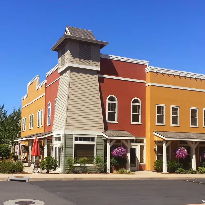 Exterior view of a colorful senior living facility building with a unique tower-like structure. The building features red, yellow, and green sections with multiple windows and hanging flower baskets. There are outdoor tables with umbrellas on the sidewalk under a clear blue sky.