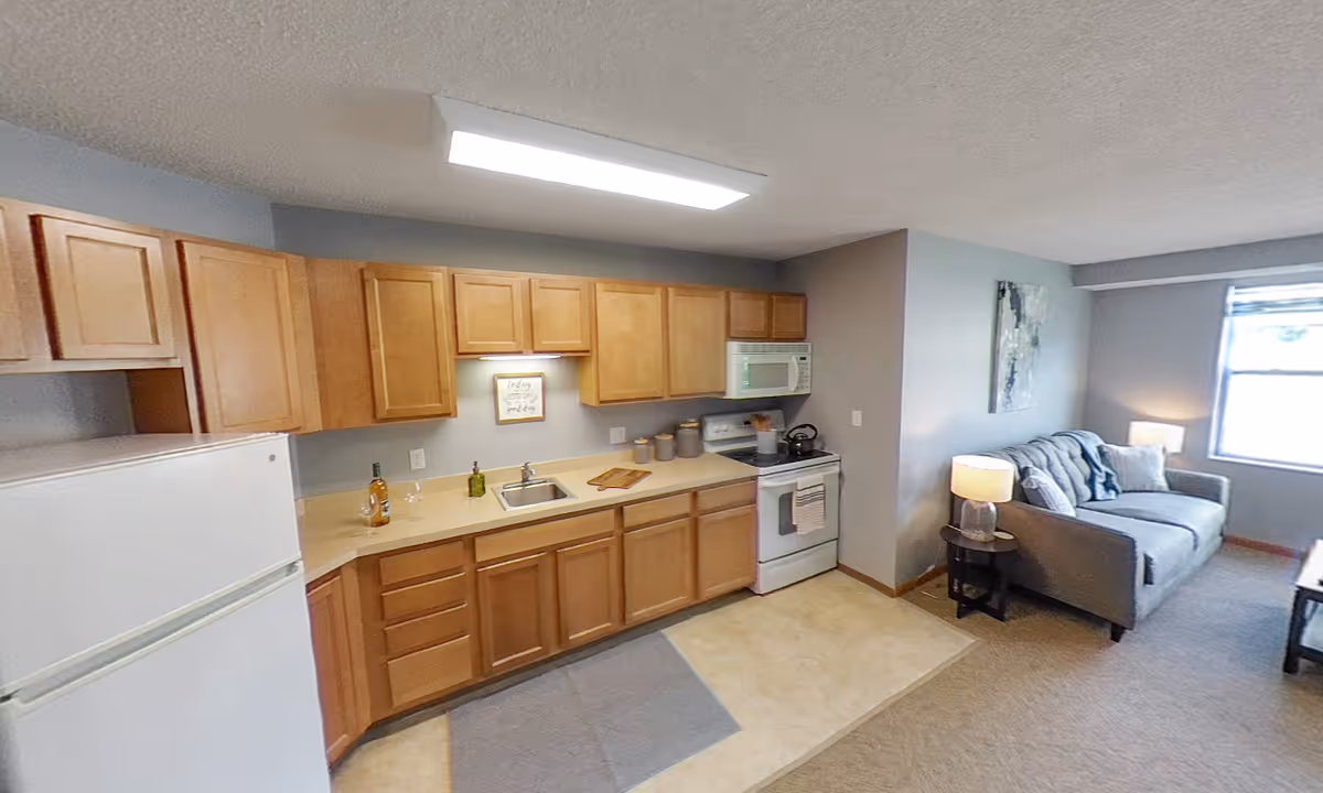 Open-plan interior showing a small kitchen with wooden cabinets, white appliances and a connected living area with a sofa and lamp.