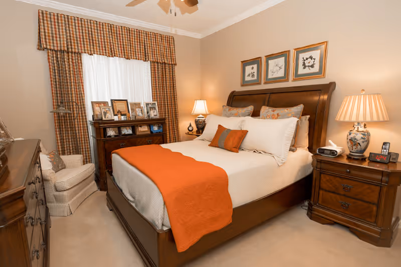 A cozy bedroom with a wooden bed frame, white bedding, and an orange throw blanket and pillows. There are two wooden nightstands with lamps on either side of the bed. A wooden dresser with framed photos and a window with plaid curtains are visible in the background. A cushioned armchair is placed near the dresser.