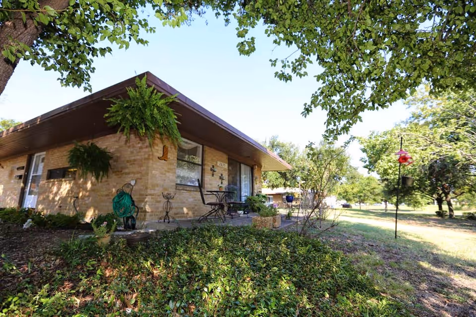 Single-story brick cottage with a covered patio, outdoor seating, hanging ferns, and a grassy yard with trees.