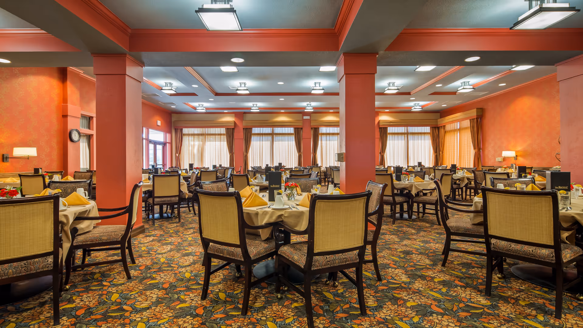 Spacious dining room with multiple tables covered in beige tablecloths and set with yellow napkins, chairs with patterned upholstery, floral carpet, large windows with sheer curtains, and warm orange walls with decorative lighting.