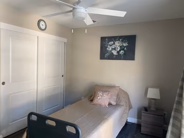 A small bedroom with a single bed covered in a beige bedspread and a pink decorative pillow. There is a nightstand with a lamp next to the bed, a floral painting on the wall above the bed, a ceiling fan, and a clock above white sliding closet doors.