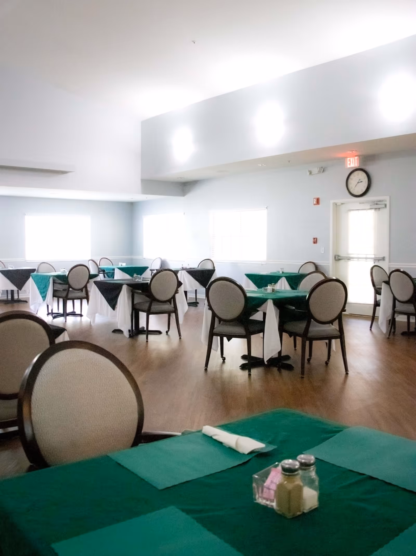 Bright dining room with round tables covered in green tablecloths and upholstered chairs on a wood floor.