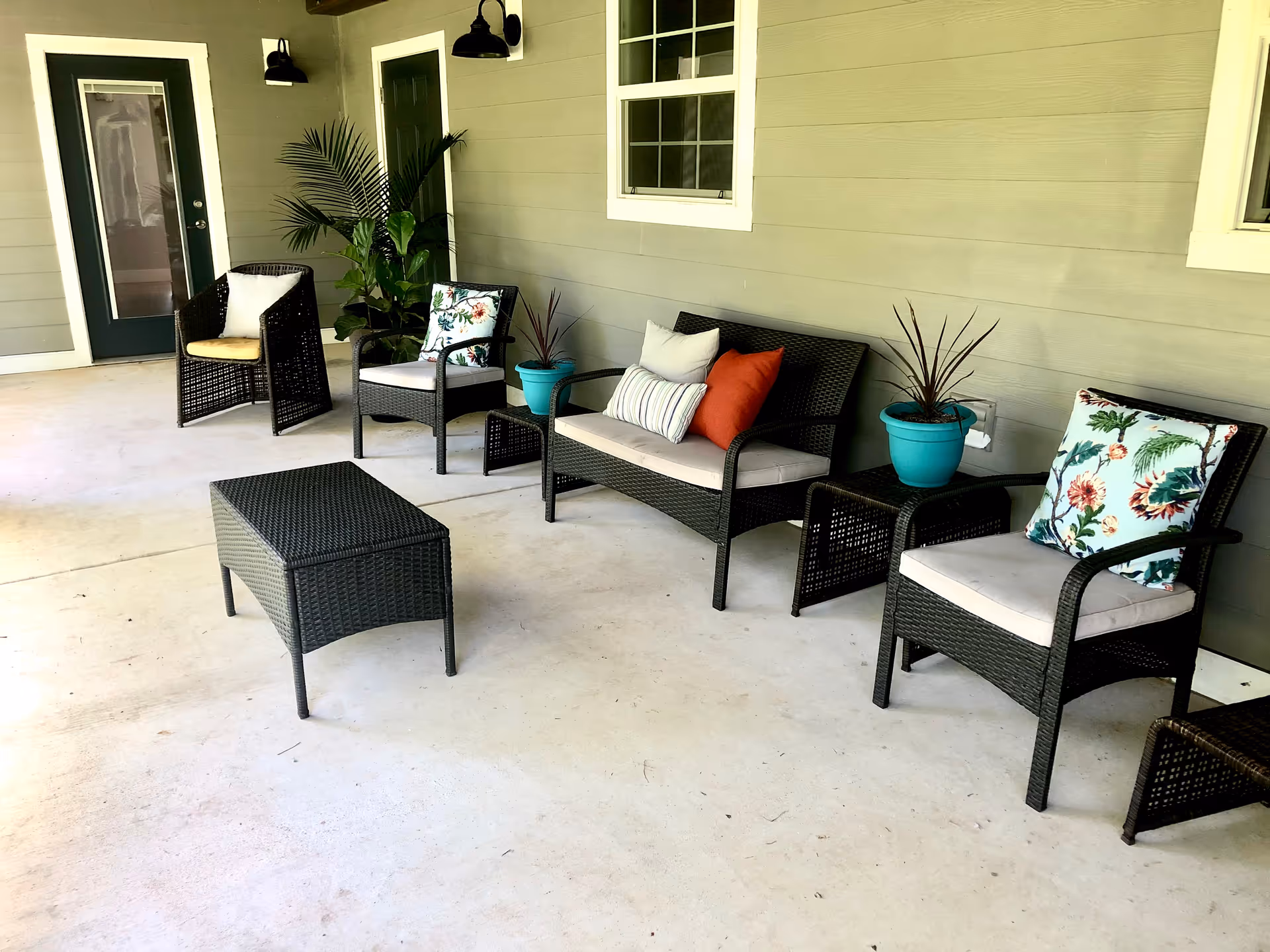 Outdoor covered patio area with wicker furniture including a loveseat, three chairs, and a rectangular coffee table. The seating has cushions and decorative pillows with floral and solid color designs. There are two potted plants on small side tables against a beige wall with two windows and a door.