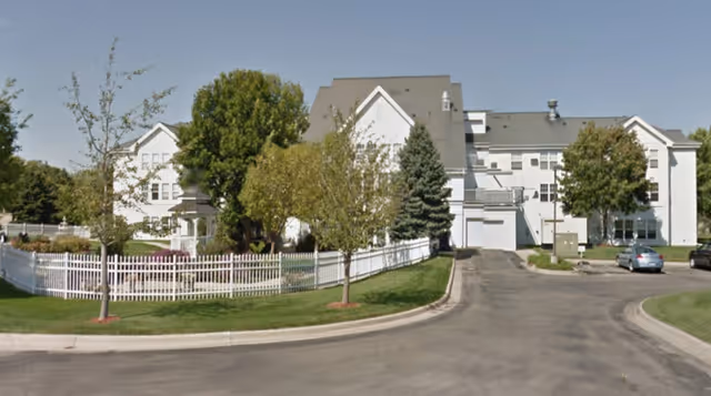 Exterior view of a large white senior living facility building with multiple windows and a gray roof. The building is surrounded by green trees, a white picket fence, and a paved driveway with parked cars.