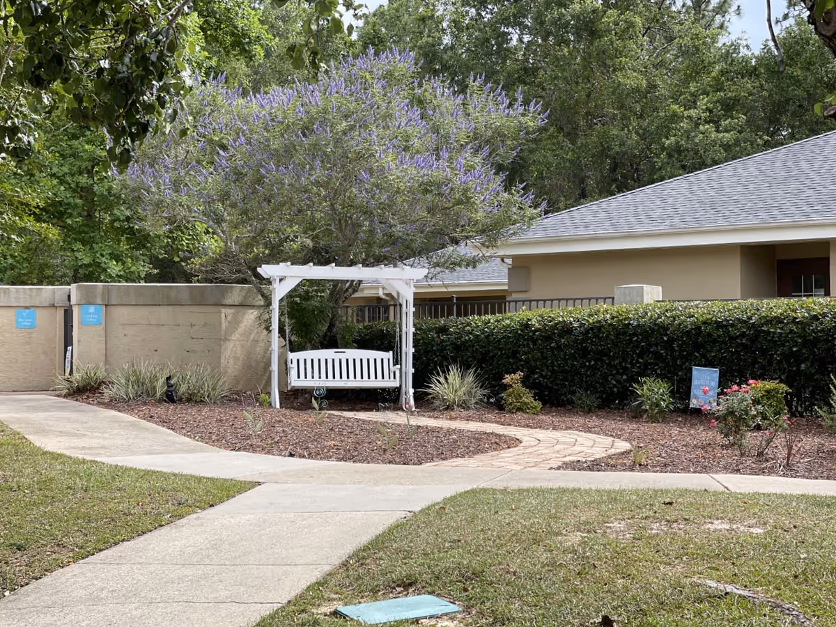 Outdoor garden area with a white wooden swing under a tree with purple flowers, surrounded by bushes and a paved walkway leading to the swing. A beige building and a concrete wall are visible in the background.