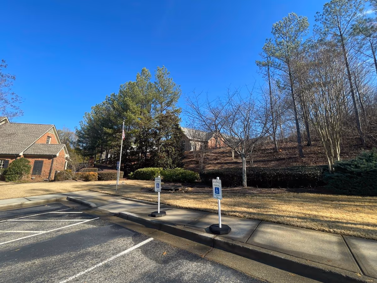 Exterior view of a senior living facility parking area with temporary handicapped signs, a flagpole, and trees under a clear blue sky.