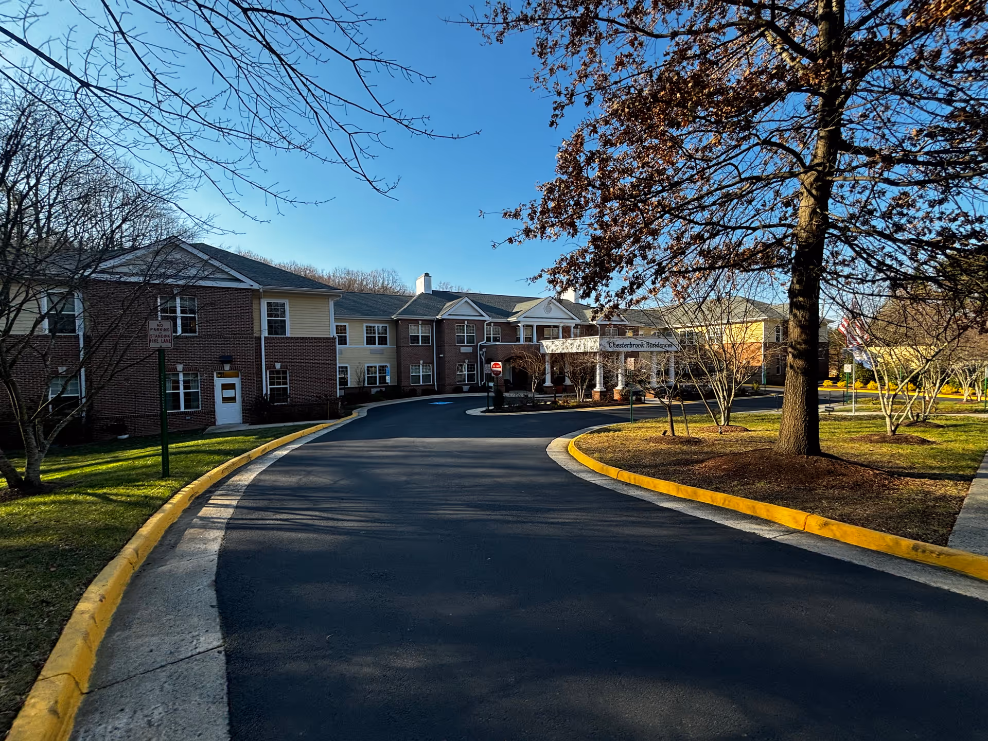 Exterior view of Chesterbrook Residences building with a curved driveway leading to the entrance. The building is two stories with a combination of brick and light-colored siding. Trees with sparse leaves and a clear blue sky are visible.