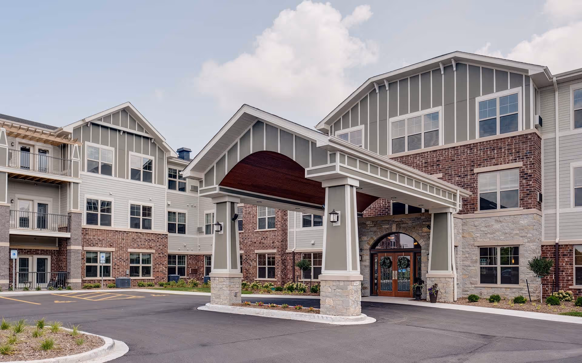 Exterior view of Aspire Senior Living Oshkosh building featuring a covered entrance with stone pillars, a combination of brick and siding facade, multiple windows, and a paved driveway with landscaping.