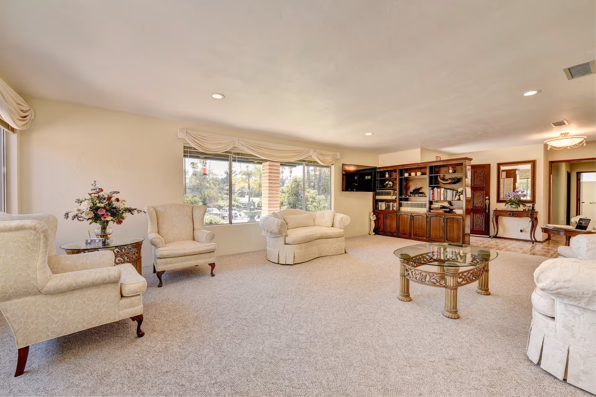 A spacious living room with beige carpet and cream-colored upholstered armchairs and loveseat. There is a glass-top coffee table with ornate legs in the center. A large window with draped valances lets in natural light and shows greenery outside. Against the far wall is a wooden bookshelf filled with books and decorative items, with a flat-screen TV mounted above it. A small table with a vase of flowers and a mirror is near the entrance hallway.