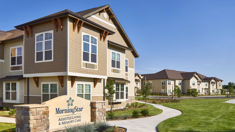 Exterior view of MorningStar Assisted Living & Memory Care facility showing multiple beige and brown buildings with large windows, a well-maintained lawn, young trees, and a curved concrete walkway under a clear blue sky.
