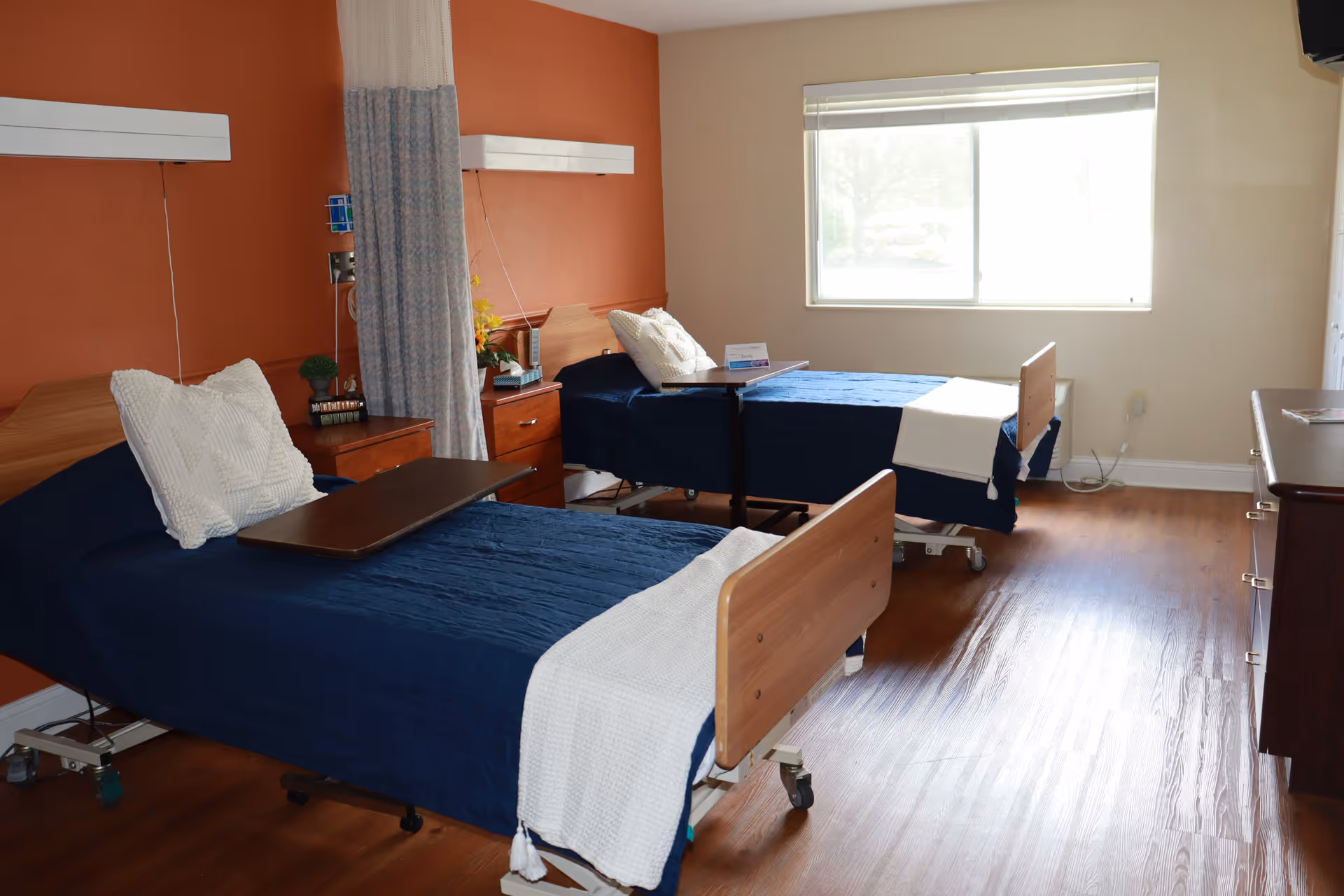 A room with two hospital-style beds with blue bedspreads and white pillows, each with a wooden headboard and footboard. There is a wooden nightstand between the beds with a small plant and other items. A curtain divider hangs between the beds. The room has wooden flooring, a large window letting in natural light, and a dresser on the right side.
