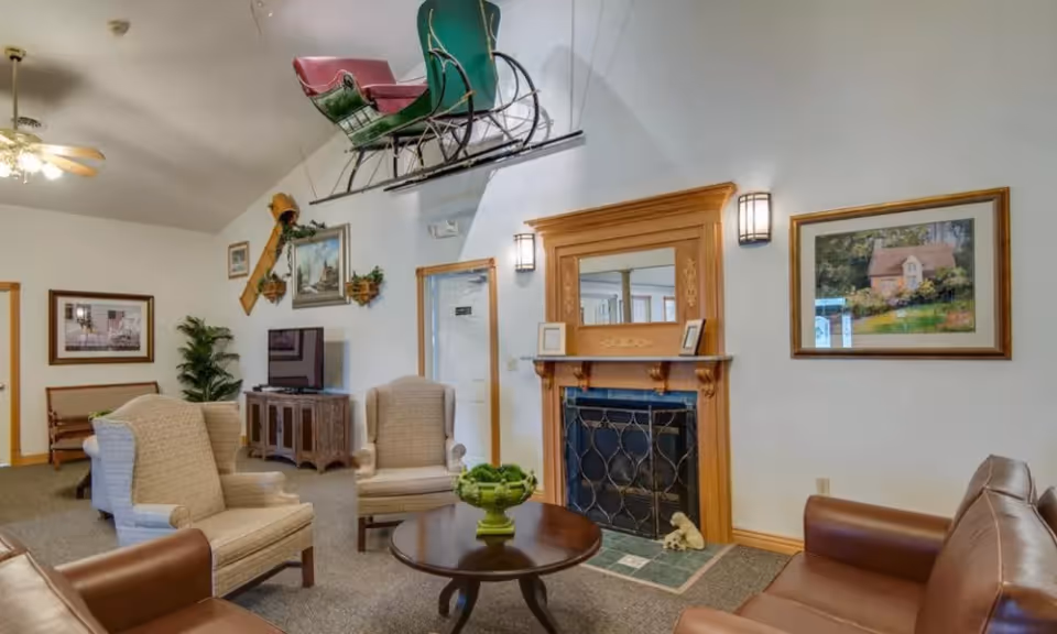 A communal living room with upholstered armchairs, a leather sofa, coffee table, wooden fireplace mantel, TV console, and decorative items including a suspended sleigh.