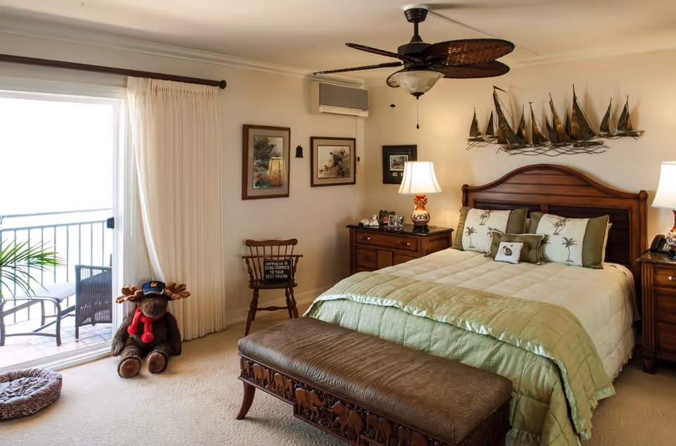 A cozy bedroom with a large bed featuring green and beige bedding and decorative pillows. There are two wooden nightstands with lamps on either side of the bed. Above the bed is a metal wall art piece shaped like sailboats. A wooden bench with carved animal designs sits at the foot of the bed. To the left, there is a sliding glass door leading to a balcony with outdoor chairs. A stuffed moose toy wearing a hat and scarf is placed near the door, next to a wooden chair with a small sign on it. The room has beige carpet and light-colored walls with framed pictures hanging.