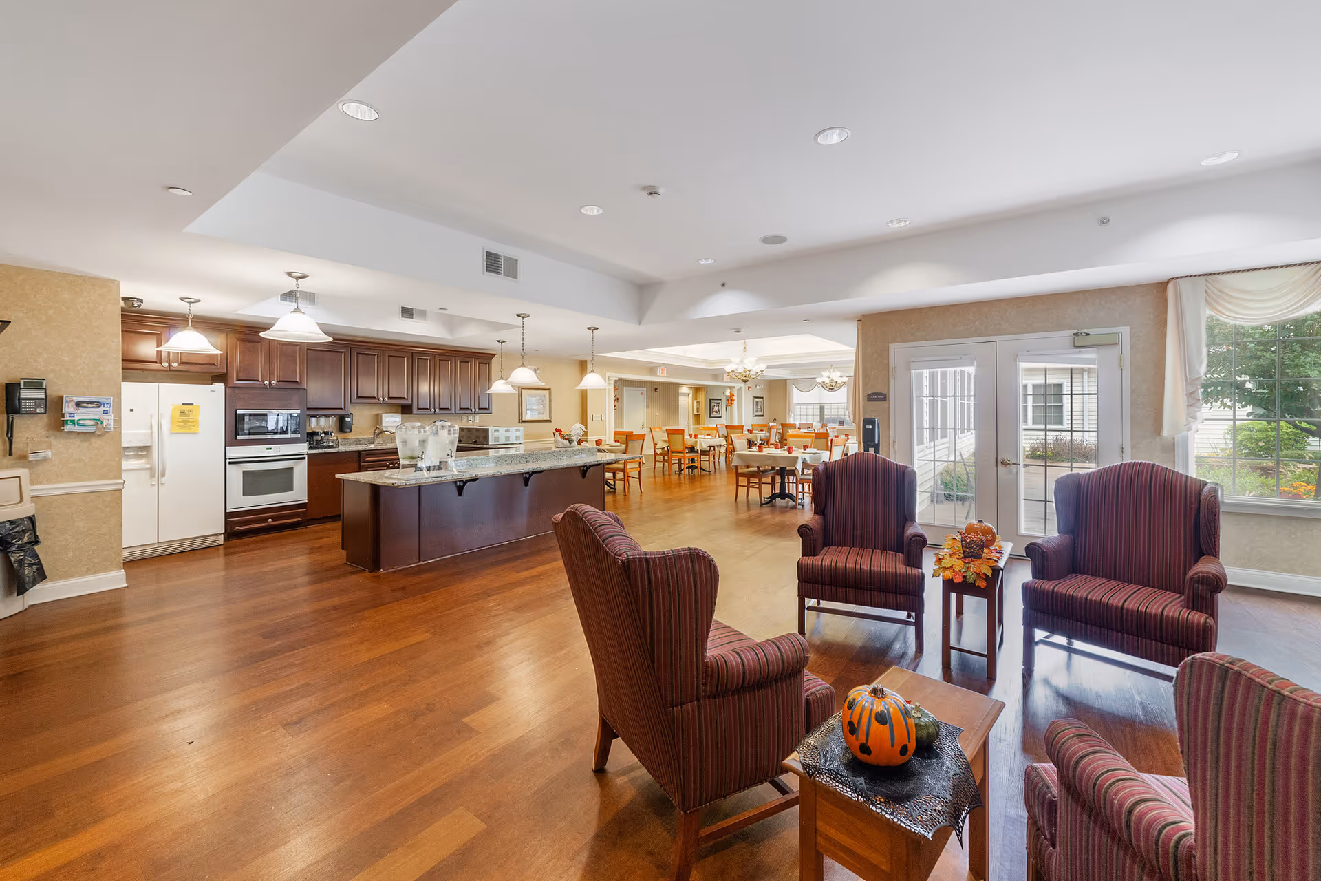 A spacious and well-lit common area in a senior living facility featuring a kitchen with dark wood cabinets, white appliances, and a large island with pendant lighting. In the foreground, there are four striped upholstered armchairs arranged around a small wooden table decorated with autumn-themed pumpkins and flowers. In the background, there is a dining area with multiple tables and chairs, large windows, and glass doors leading outside.