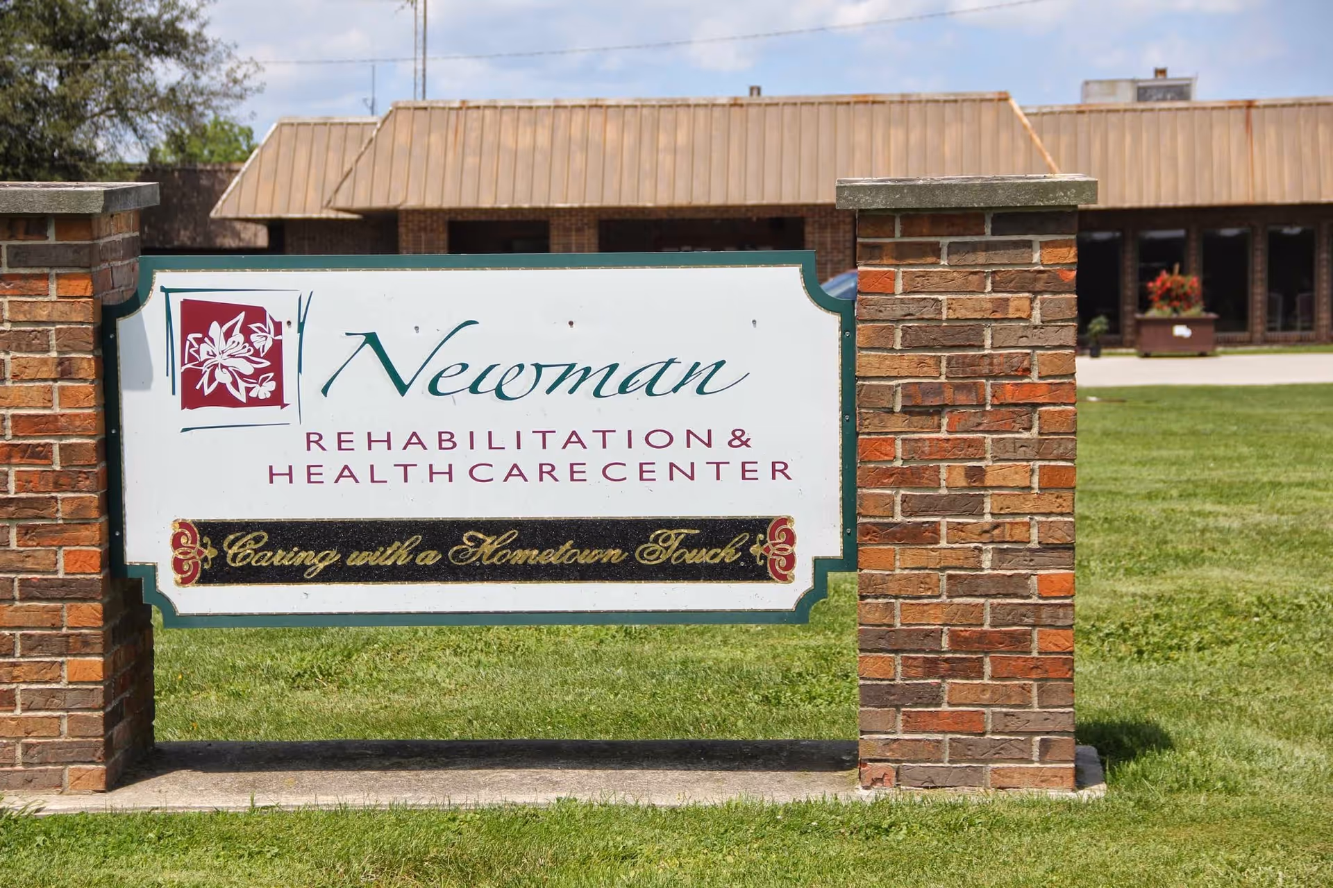 Outdoor view of a sign for Newman Rehabilitation & Health Care Center mounted between two brick pillars with a building and green lawn in the background.