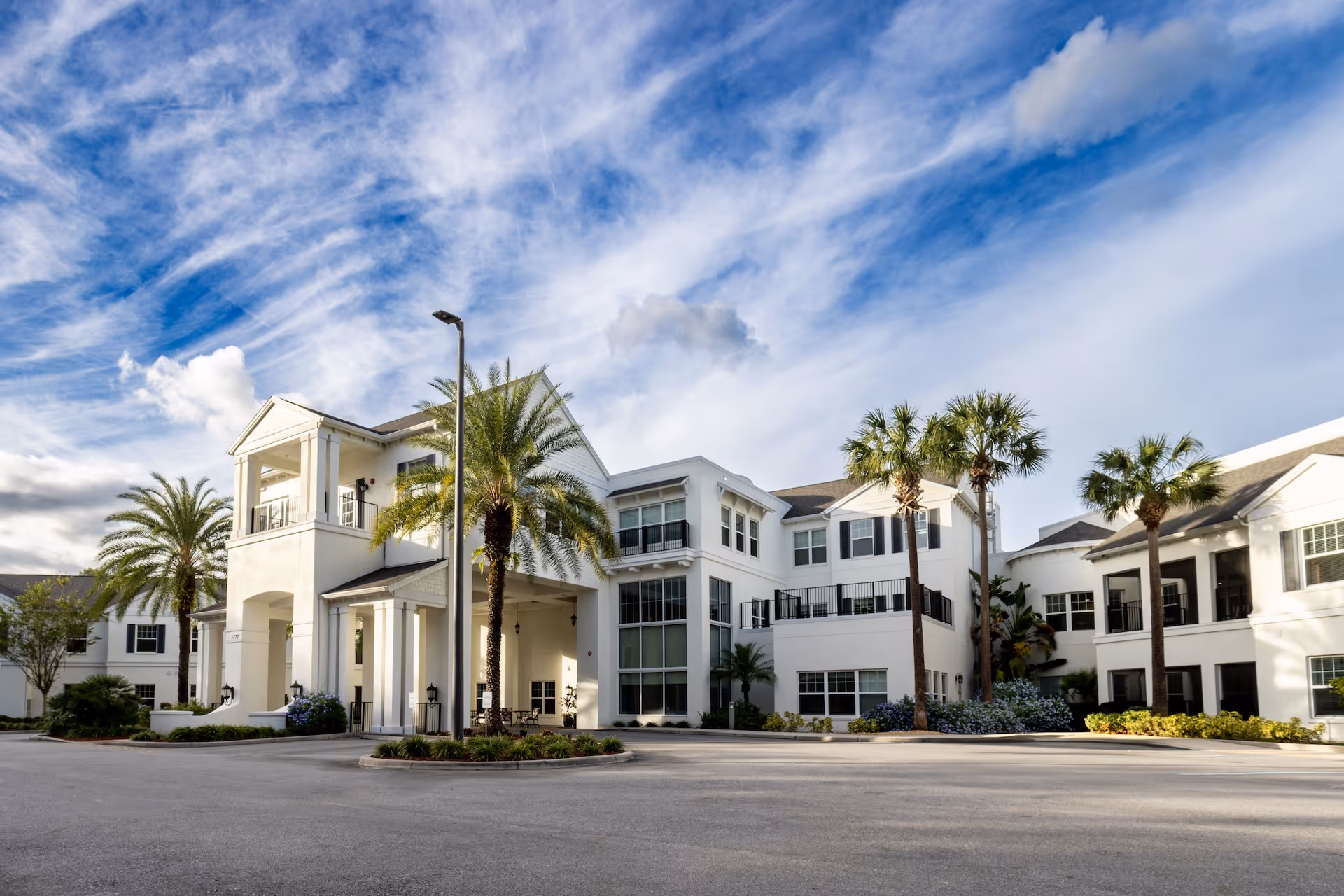 Exterior view of a large, white senior living facility building with multiple windows and balconies, surrounded by palm trees and landscaping under a partly cloudy blue sky.