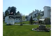 A large white multi-story building with multiple windows and a sloped roof, set behind a well-maintained green lawn. In the foreground, there is a white gazebo on the left and a stone well structure on the right. Trees and shrubs are scattered around the lawn under a clear blue sky.
