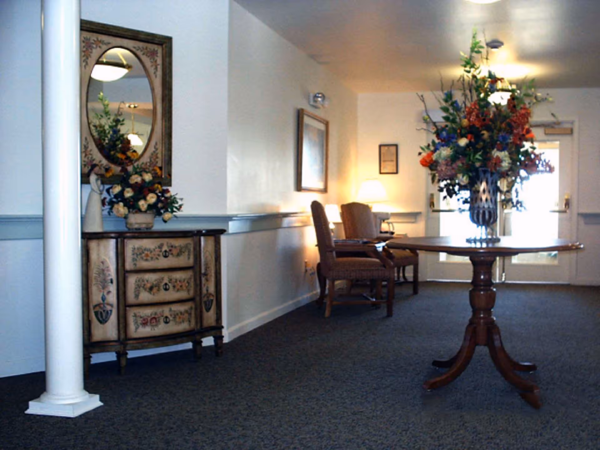 Interior hallway area with a decorative wooden table holding a large floral arrangement in a vase. To the left, there is a small painted chest of drawers with a matching framed mirror above it and a smaller floral arrangement on top. Two upholstered chairs and a lamp are positioned against the wall near a door with glass panels letting in natural light.
