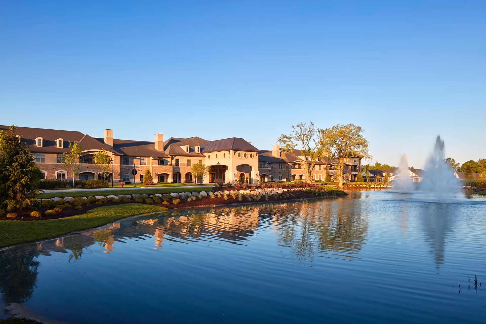 A large senior living facility building with a beige and brown exterior, situated beside a pond with a water fountain. The building is surrounded by landscaped gardens and trees under a clear blue sky.