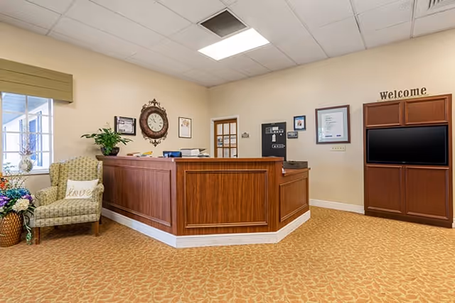 Reception area with a wooden front desk, a patterned armchair with a pillow that says 'Love', a large wall clock, framed pictures on the wall, a TV mounted in a wooden cabinet with a 'Welcome' sign above it, and a window with a green valance.
