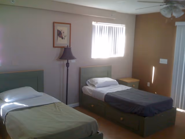 Two single beds in a simple, well-lit bedroom with a floor lamp, nightstand, and window blinds.