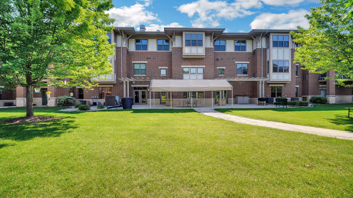 A three-story brick building with multiple windows and a covered patio area in the center. The building is surrounded by a well-maintained green lawn with a paved walkway leading to the patio. There are two large trees on either side of the lawn under a partly cloudy blue sky.