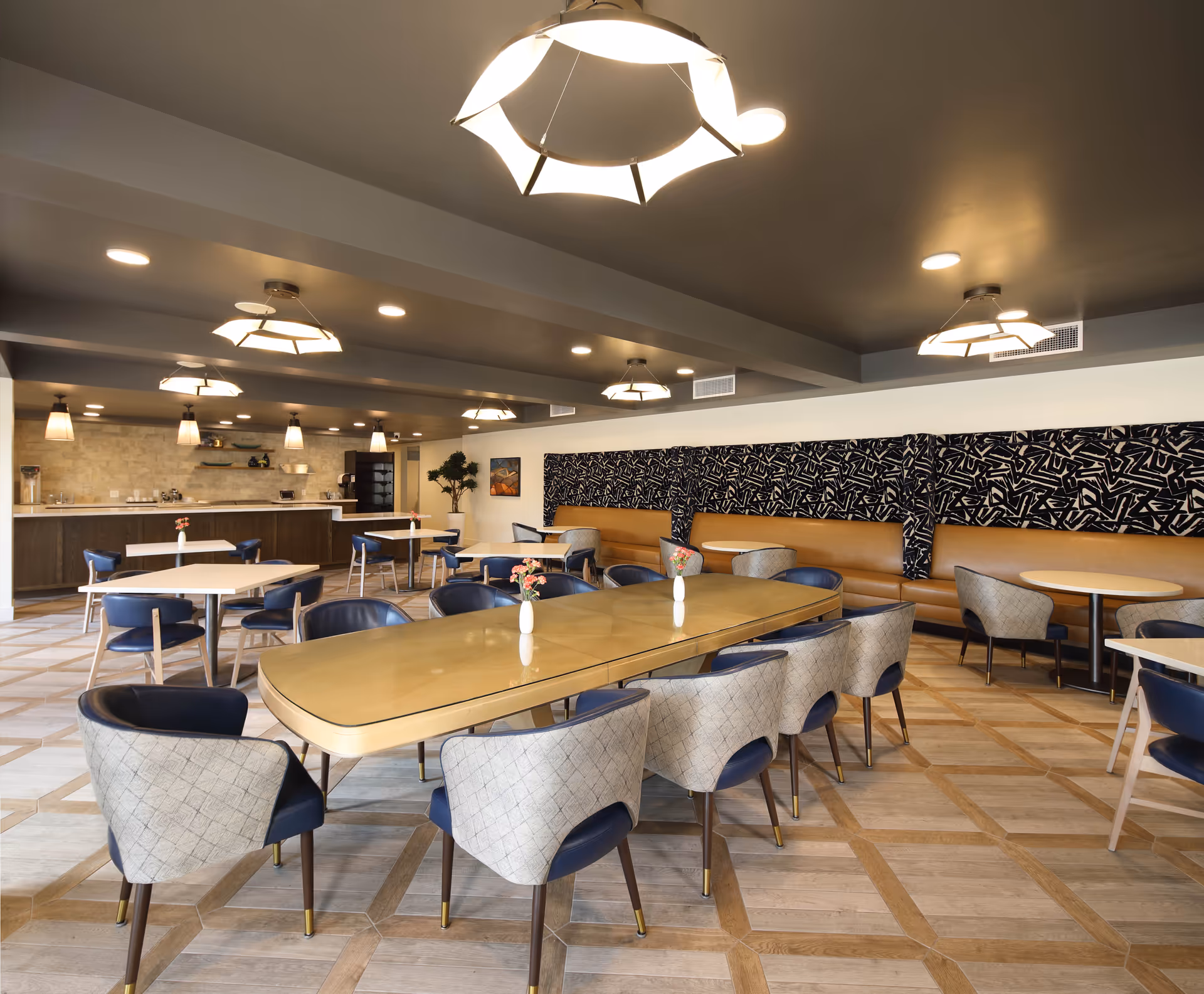 A modern dining room in a senior living facility with multiple tables and chairs arranged neatly. The room features a long table with cushioned chairs in the foreground, smaller tables with blue chairs, a cushioned bench along the wall with patterned upholstery above it, and a counter area with pendant lights and shelves in the background. The floor has a geometric wood pattern, and the ceiling has recessed lighting and contemporary hanging light fixtures.