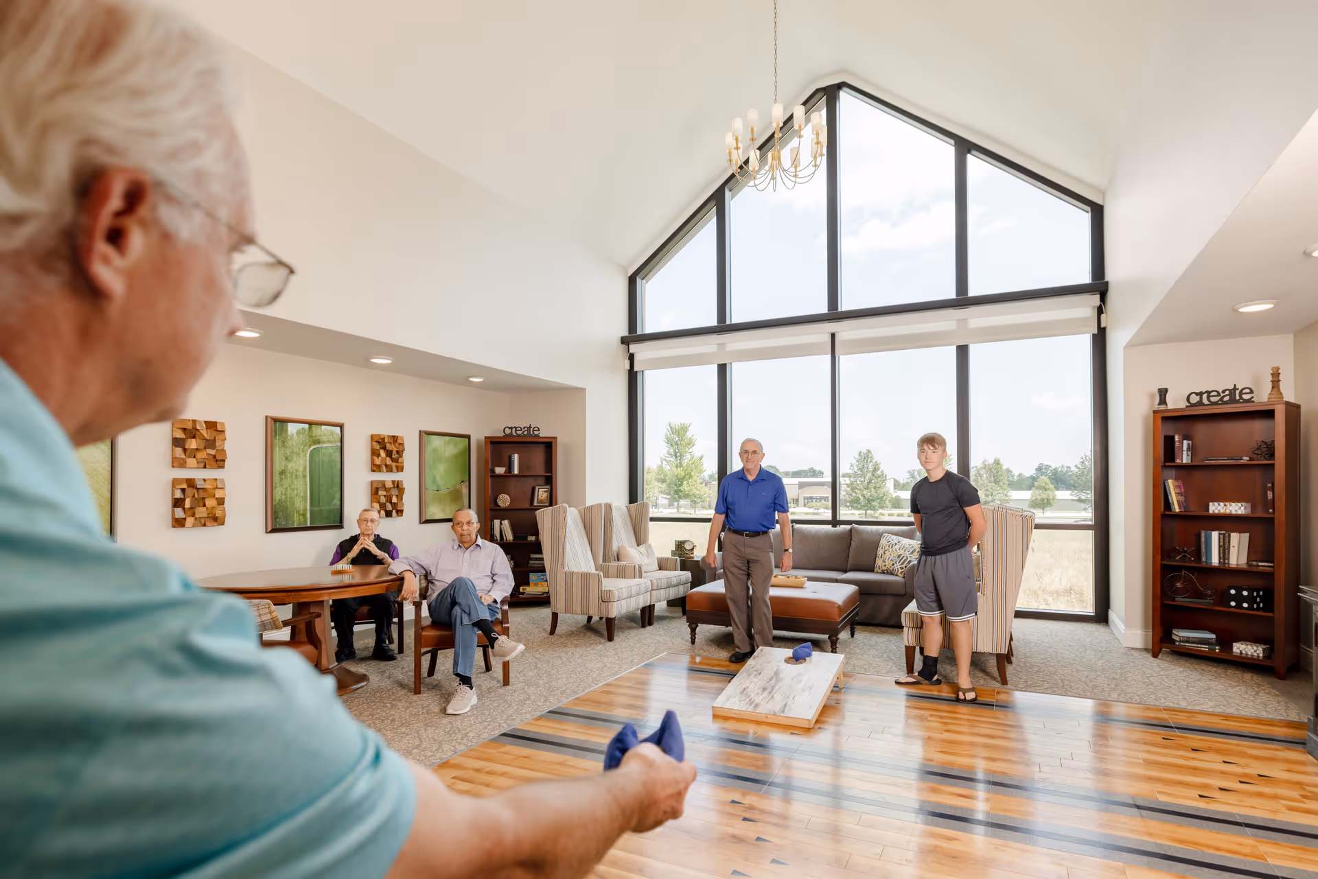 A group of four men in a bright living room with large windows and high ceilings. One man in the foreground is playing a game with a wooden board on the floor, while two men sit at a table and two others stand near a couch and armchairs. The room has wooden floors, carpet, bookshelves, and wall art.