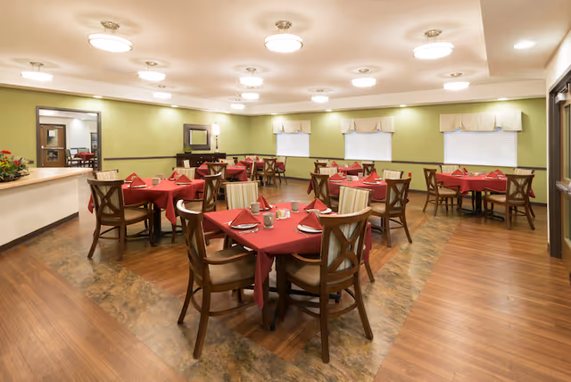 A dining room with multiple tables covered in red tablecloths, each set with napkins, glasses, and silverware. The room has wooden floors, light green walls, and several windows with valances. Ceiling lights illuminate the space evenly.
