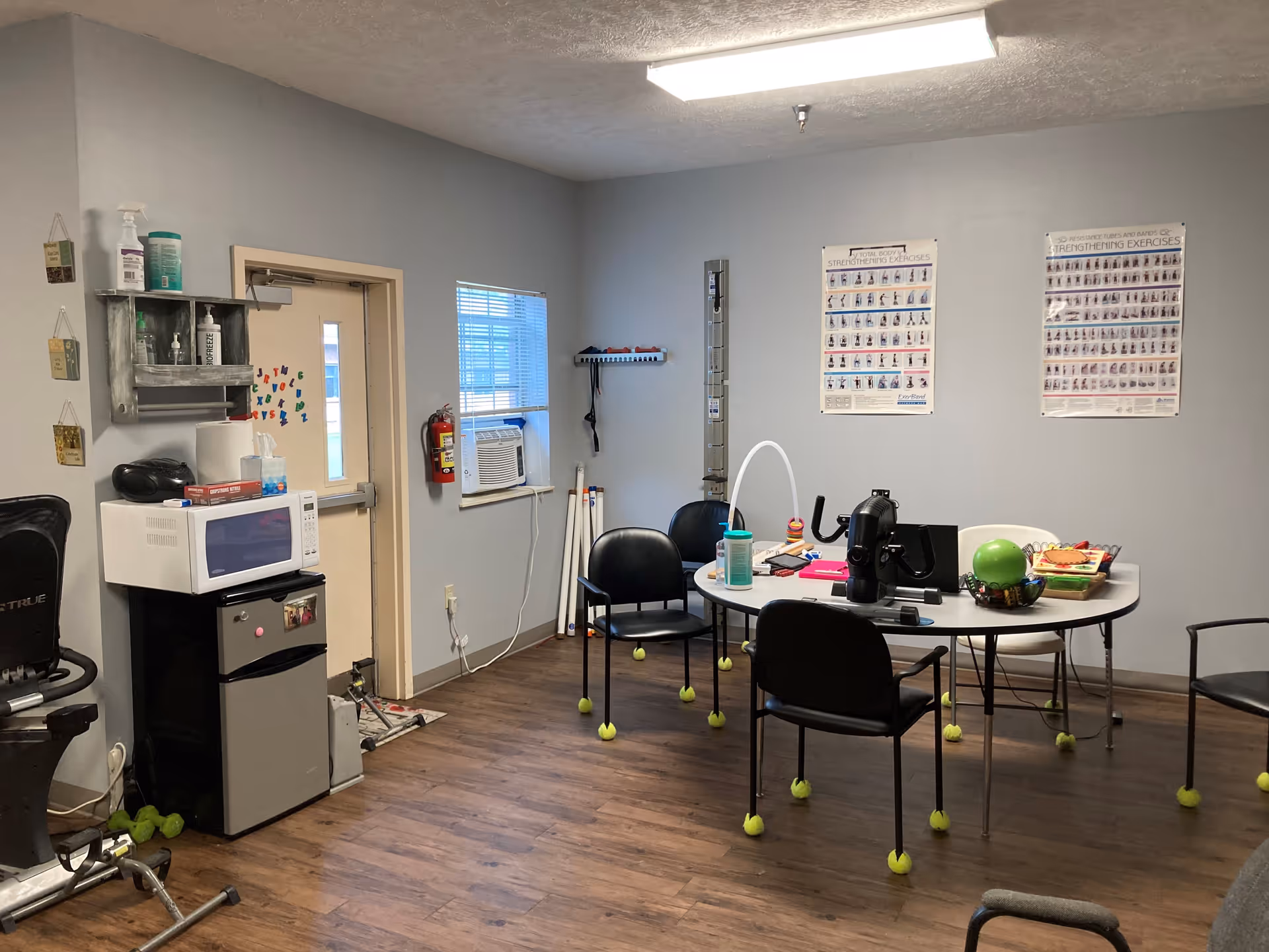 A room with exercise and therapy equipment including a stationary bike, a table with various items, and chairs with tennis balls on the legs. There is a microwave and mini fridge against one wall, a window with an air conditioning unit, and two posters on the wall showing strengthening exercises.