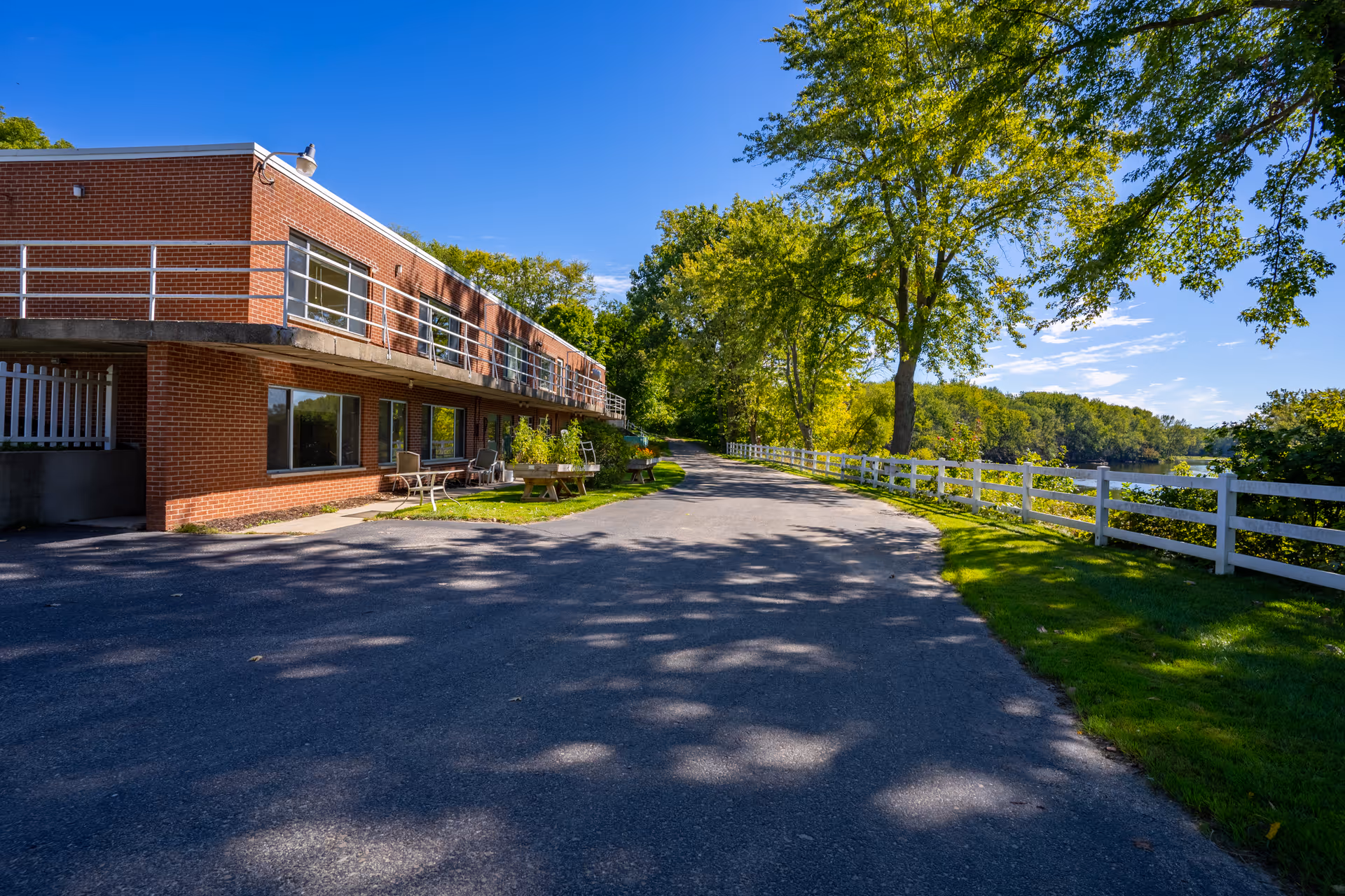 A two-story brick building with outdoor seating on the ground level, situated next to a paved driveway. There are large trees and a white fence lining the right side of the driveway, with a body of water visible beyond the fence under a clear blue sky.
