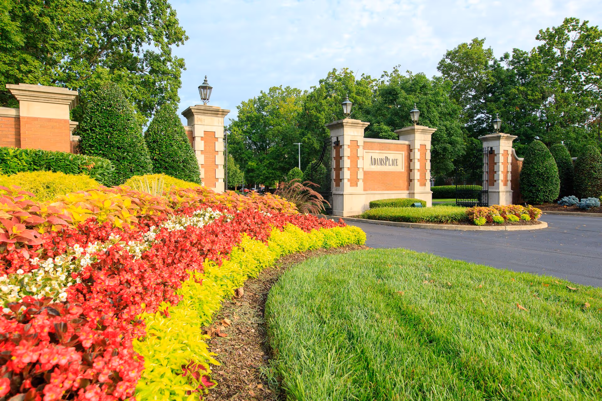 Entrance to AdamsPlace facility with a brick and stone gate, surrounded by neatly trimmed bushes and colorful flower beds, under a partly cloudy sky.