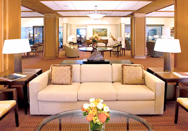 A spacious and well-lit living room area with a beige sofa adorned with two patterned cushions, flanked by two side tables each holding a lamp. In front of the sofa is a glass coffee table with a floral arrangement. The background shows additional seating areas, wooden columns, and a piano, creating a warm and inviting communal space.