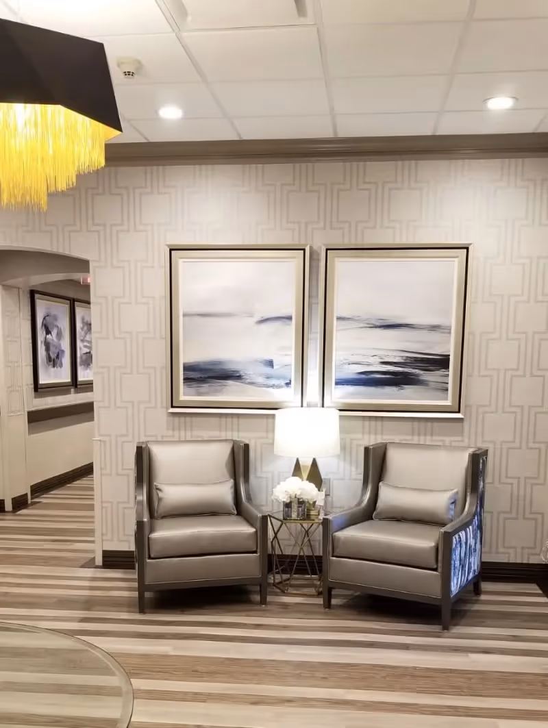 A cozy seating area with two gray armchairs separated by a small round table holding a lamp and a flower arrangement. Behind the chairs are two abstract paintings with blue and white tones on a patterned beige wall. The floor has light wood stripes, and a hallway with additional artwork is visible to the left.