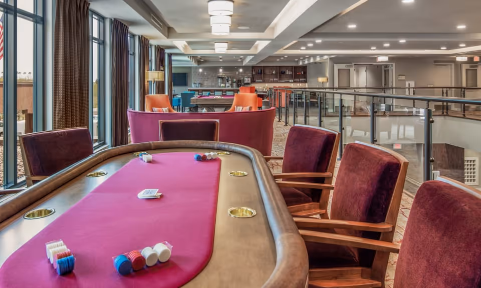 Interior view of a senior living facility's recreational area featuring a poker table with chips and cards, surrounded by purple upholstered chairs. The space includes large windows with curtains, modern lighting fixtures, and a bar area with additional seating in the background.