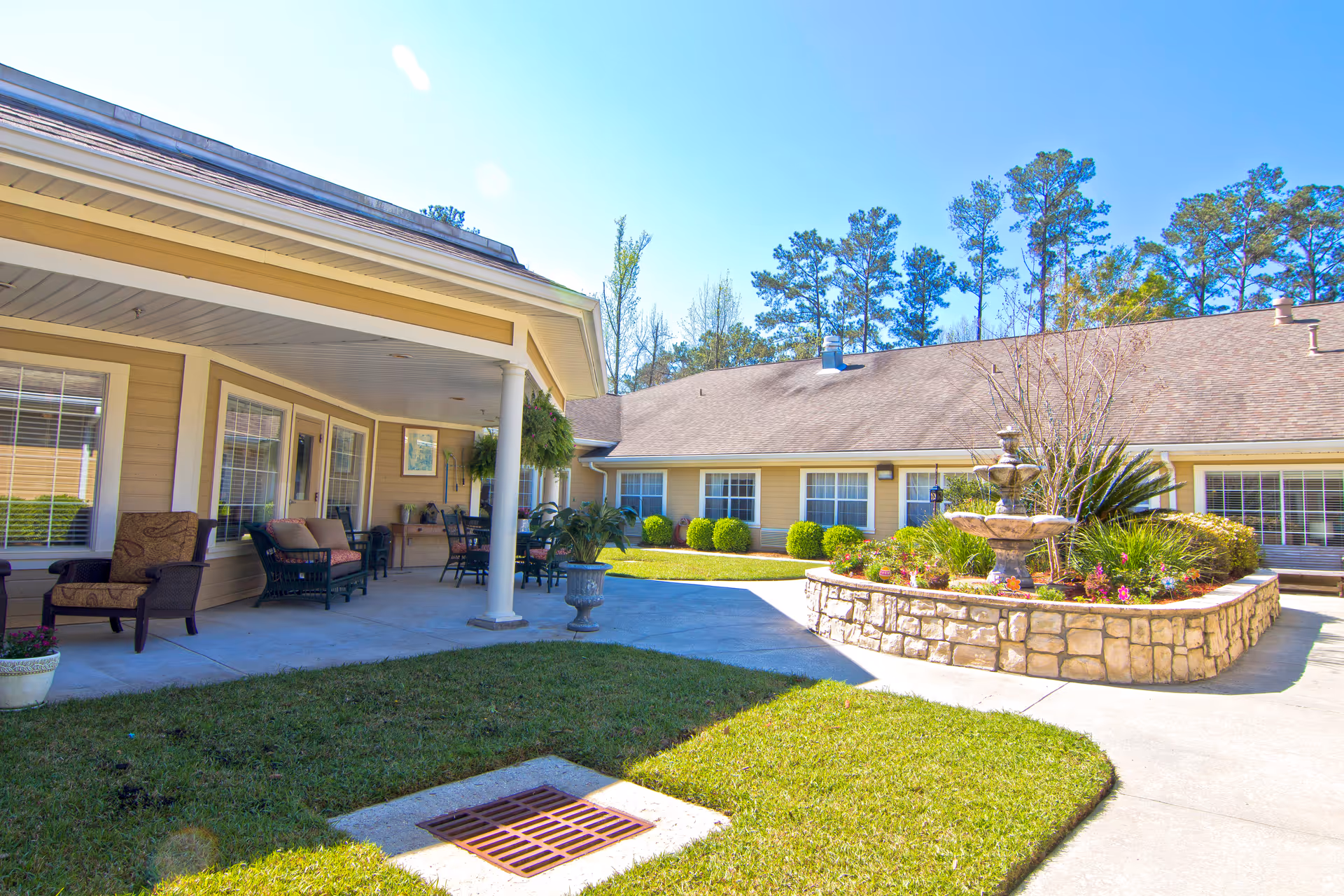 Outdoor courtyard area of a senior living facility with a covered patio featuring chairs and tables, a stone planter with a multi-tiered fountain surrounded by flowers and greenery, and a building with multiple windows in the background under a clear blue sky.