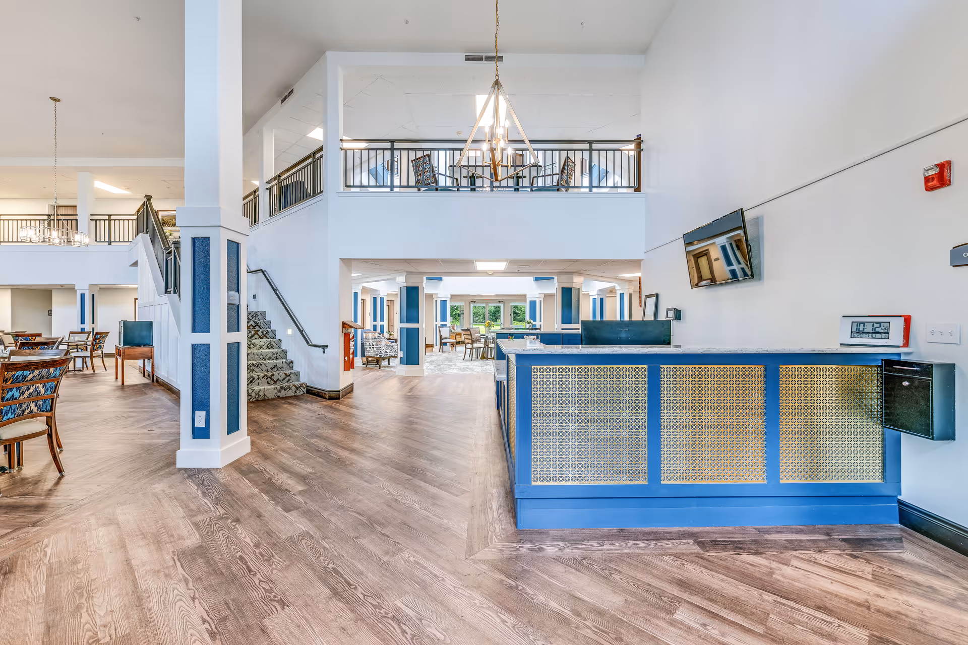Bright open senior living lobby with a reception desk, seating and dining areas, and a staircase to an upper level.