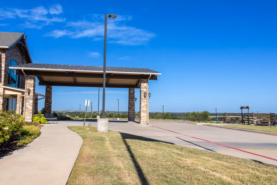 Exterior view of Bristol Park at Cleburne Assisted Living & Memory Care showing a covered entrance with stone pillars, a sidewalk, a driveway, and a clear blue sky in the background.