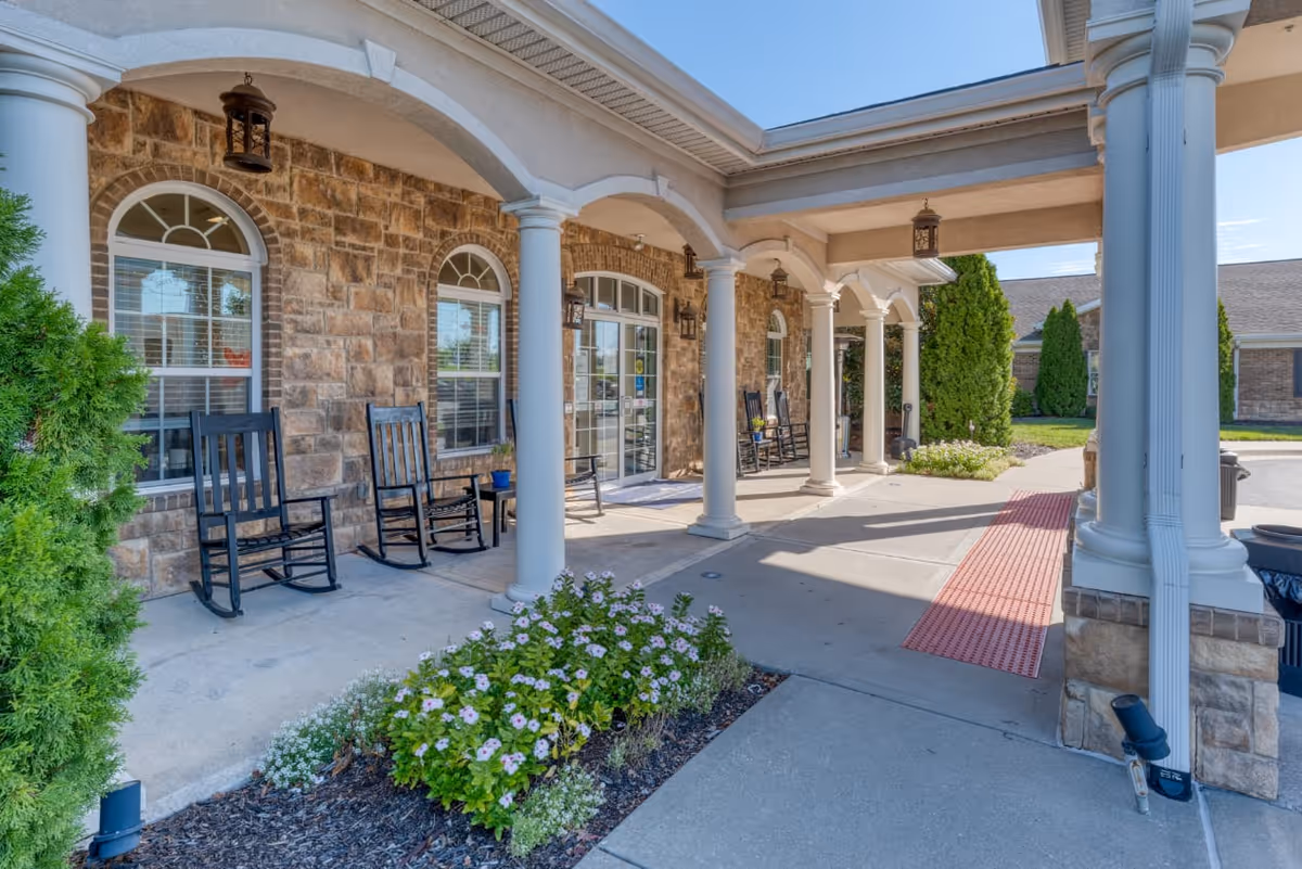 Covered entrance area of a building with stone walls and white columns. There are black rocking chairs and small tables along the wall under the covered porch. Flower beds with blooming plants are in front of the porch, and a red textured mat is on the concrete walkway leading to the entrance door.