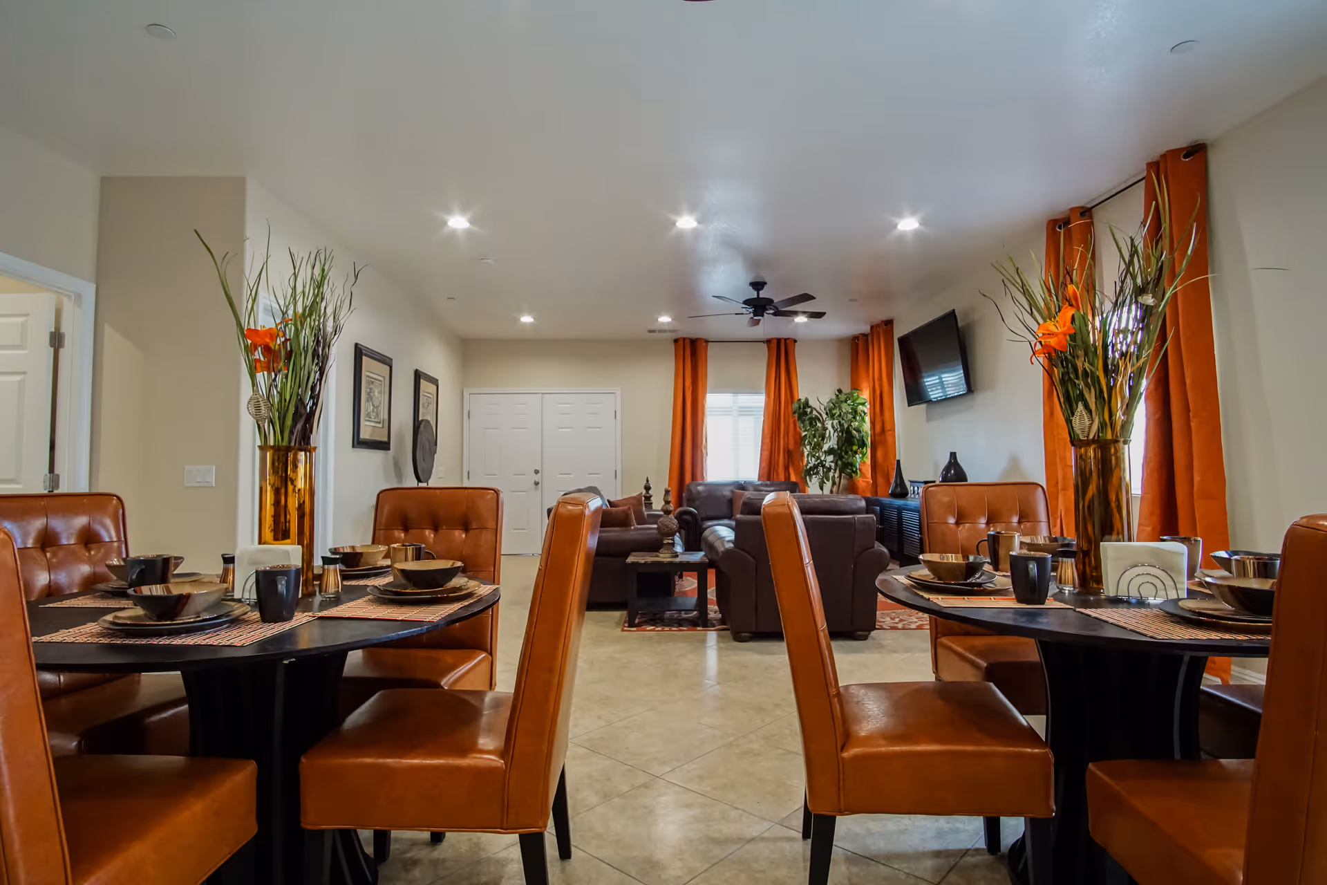Interior view of a senior living facility dining and living area with two round dining tables set with dishes and cups, surrounded by brown leather chairs. The living area in the background has brown leather sofas, a wall-mounted TV, orange curtains, and decorative plants.