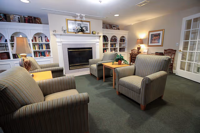 A cozy living room with four striped armchairs arranged around two wooden side tables. There is a fireplace with a white mantel in the center of the back wall, flanked by built-in white bookshelves filled with books and decorative items. A table lamp is on a side table to the left, and a framed picture hangs above the fireplace. The room has carpeted flooring and soft lighting.
