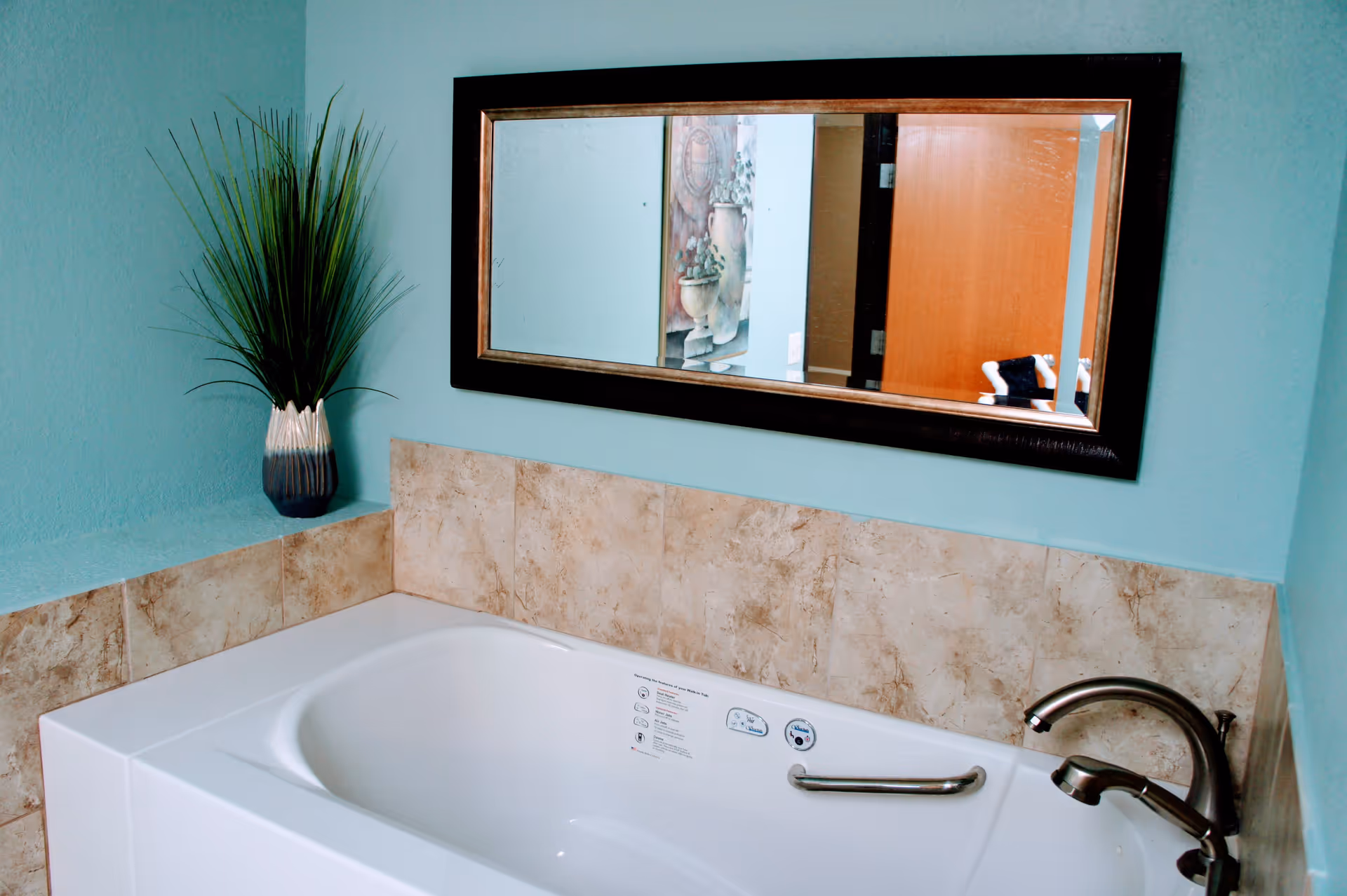 A white bathtub with a tiled surround, metal faucet and grab bar beneath a rectangular mirror on teal walls with a potted plant.