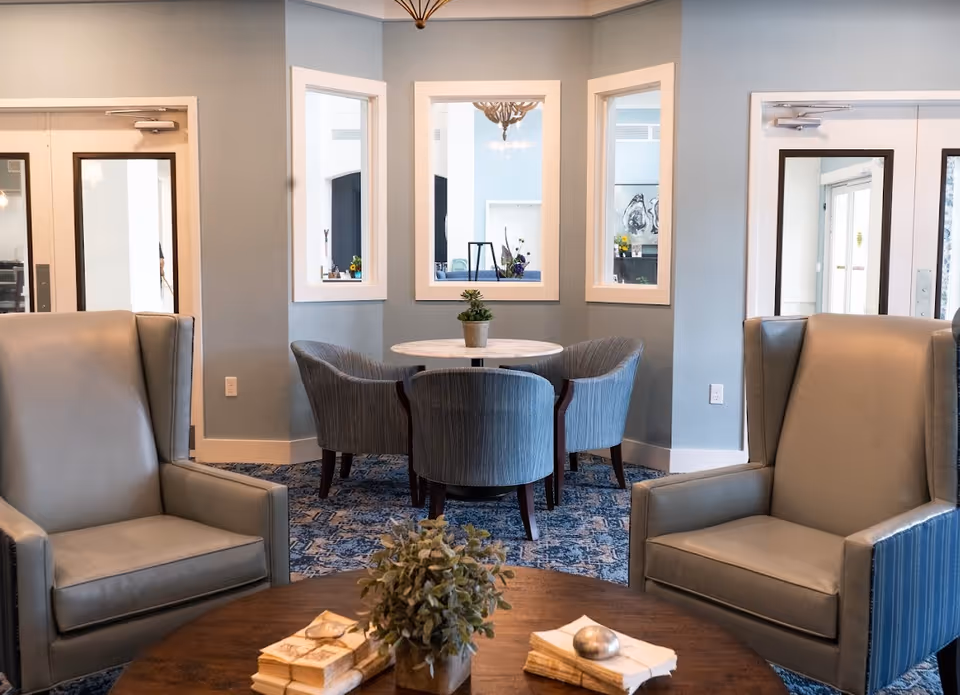A cozy seating area in a senior living facility with two gray armchairs facing a round wooden table that has a small plant and some books on it. In the background, there is a round table with four gray upholstered chairs, set against a light blue wall with three windows showing another room.