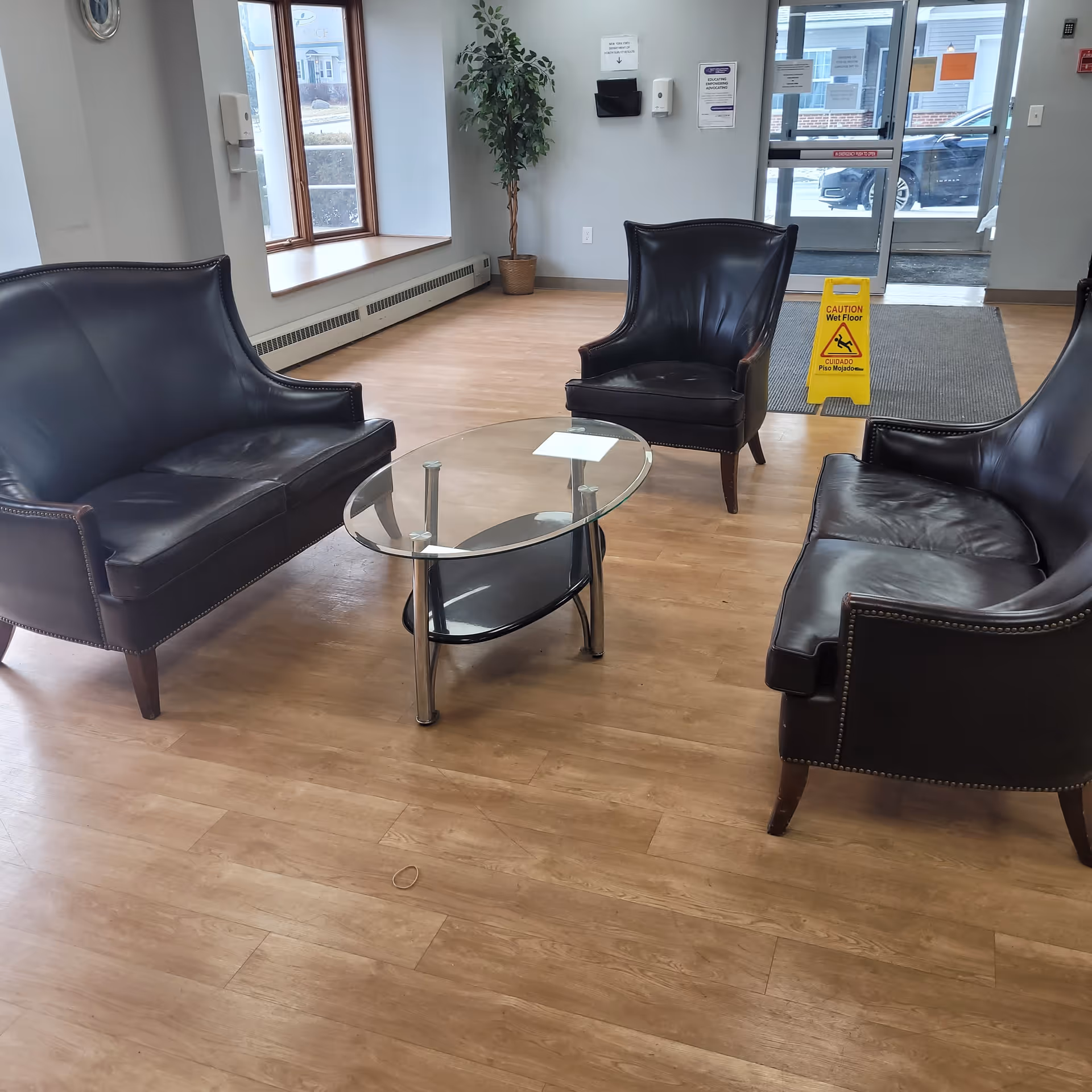 Lobby seating area with three dark leather chairs and a loveseat around a glass coffee table on wood flooring near an exterior door and a yellow wet-floor sign.