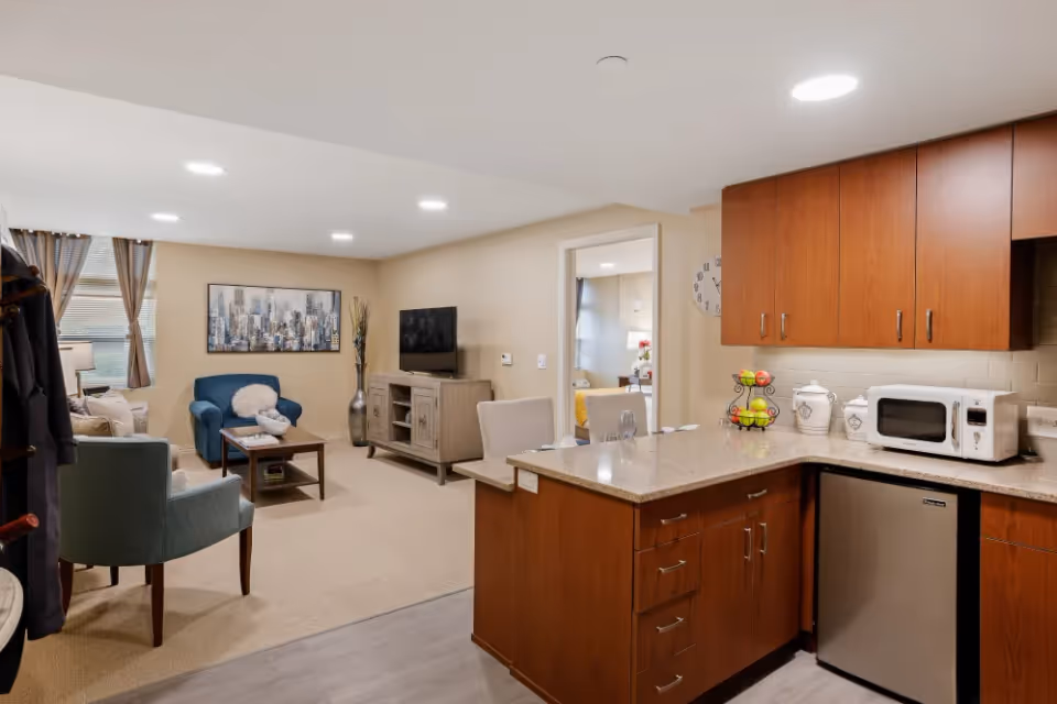 Interior view of a senior living facility showing a combined living room and kitchen area. The living room has a blue armchair, a beige sofa, a wooden coffee table, a TV on a wooden stand, and a large window with curtains. The kitchen features wooden cabinets, a granite countertop, a microwave, a small refrigerator, and a fruit basket. A doorway leads to a bedroom with a bed and a lamp visible.