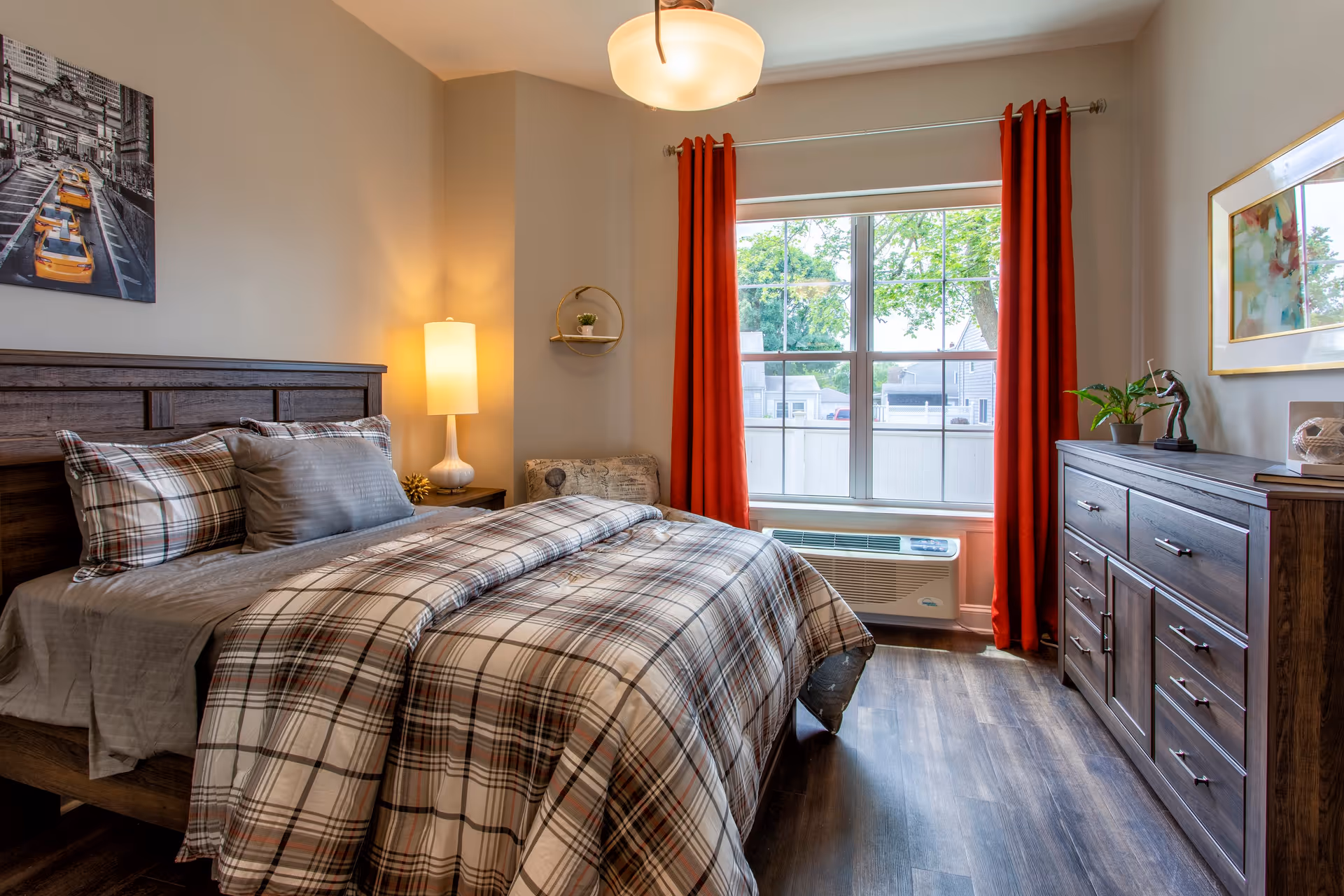 Sunlit bedroom with a plaid bed, wooden headboard and dresser, red curtains, and a window air conditioner.