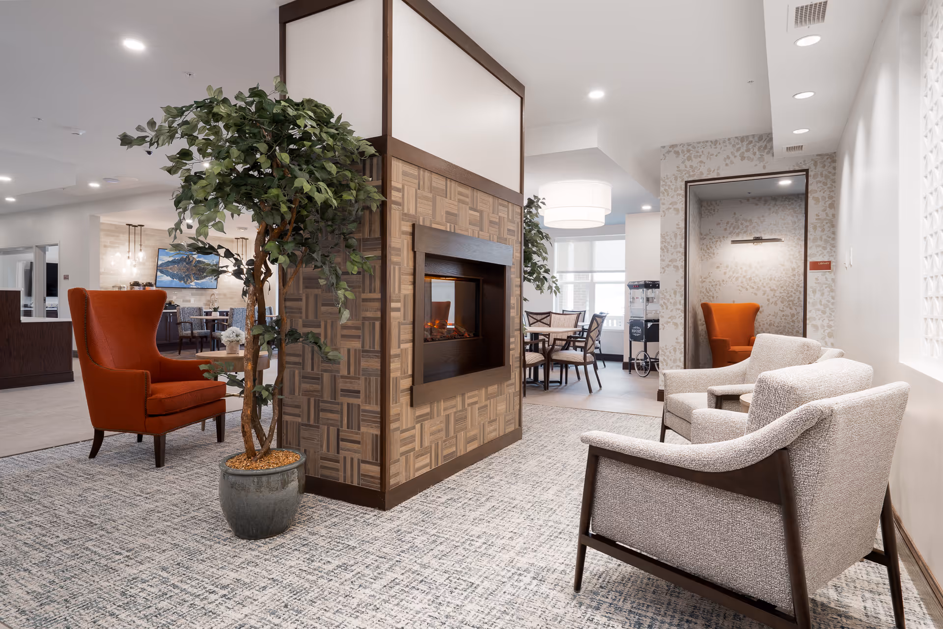 A bright communal lounge with armchairs, a central decorative fireplace, a potted tree, and a dining area visible in the background.